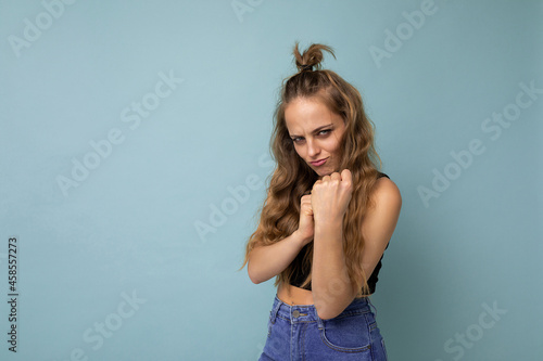 Young beautiful blonde wavy-haired woman with sincere emotions wearing trendy black top isolated over blue background with empty space and ready to fight