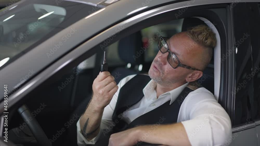 Happy car owner admiring key turning to camera with surprised facial expression. Portrait of handsome Caucasian lottery winner posing sitting on driver's seat in new luxurious vehicle in dealership Happy car owner admiring key turning to camera with surprised facial expression. Portrait of handsome Caucasian lottery winner posing sitting on driver's seat in new luxurious vehicle in dealership