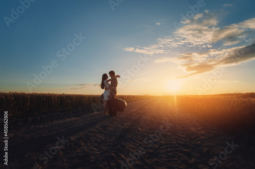 Young lovers caucasian newlyweds stand and hug before a kiss against the background of the sunset. Silhouette of a guy with a guitar and a girl on the road between wheat fields.Country style.Wild west