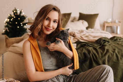 A pretty smiling young woman in pajamas hugs her friend's pet domestic cat sitting in a cozy bedroom decorated with Christmas decor at home, selective focus