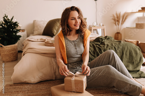 A smiling happy young woman in pajamas holds a Christmas gift box sitting on the floor by the bed in a cozy decorated room at home during the winter holidays. Selective focus