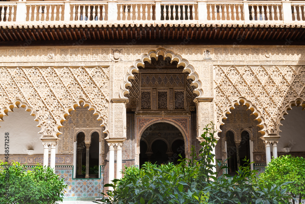 Fototapeta premium The arab style decorated facade in a patio of the famous Alcazar (meaning: fortress) in Seville, Andalusia, Spain. Green plants in the foreground.