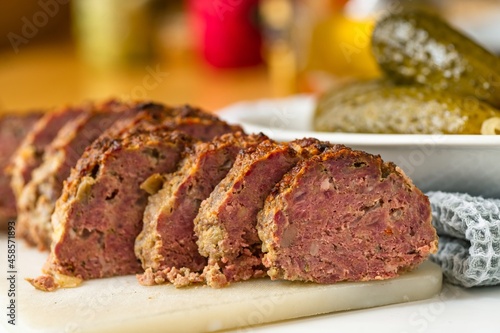 Baked sliced beef meatloaf, pickled cucumber and towel on kitchen table.