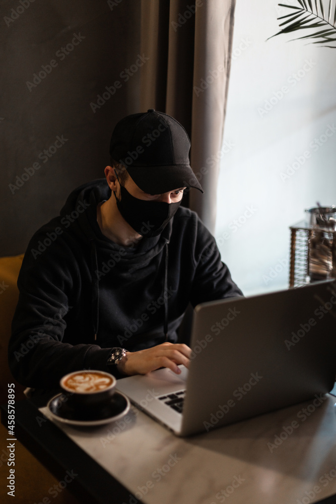 Fashionable man in stylish black clothes with a mockup and a black protective mask is sitting in a cafe, drinking coffee and working on laptop Fashionable man in stylish black clothes with a mockup and a black protective mask is sitting in a cafe, drinking coffee and working on laptop