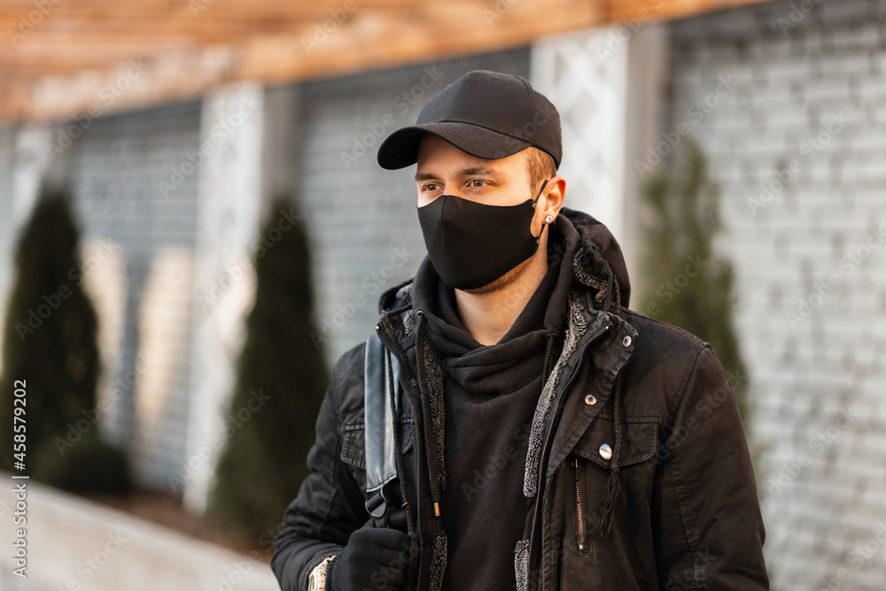 Young man with a black protective mask and a fashionable cap in a black stylish hoodie and a winter jacket with a backpack walks in the city