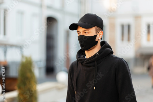 Stylish young man in black mock up clothes with cap, hoodie and protective covid mask walks in the city