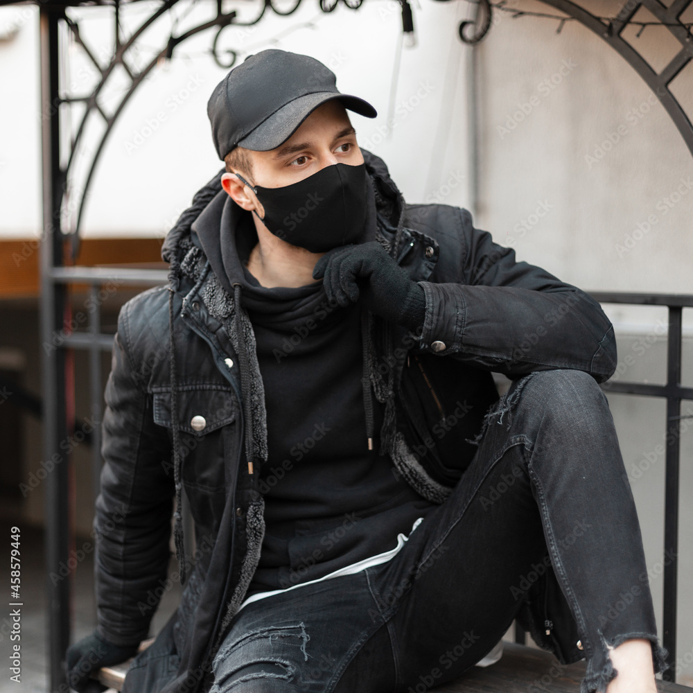 Fashionable handsome hipster guy with a black stylish cap and a protective medical mask in a black jacket and a hoodie sits in the city. Urban male casual modern style