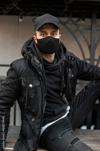 Handsome young man model with black cap and protective medical mask in black jacket and hoodie sits on the street