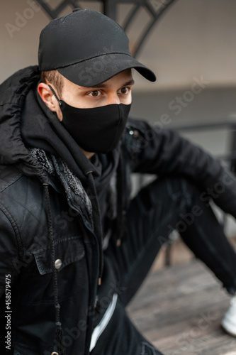 Fashionable handsome young man with protective black mask in fashion black jacket and hoodie with cap sits on the street