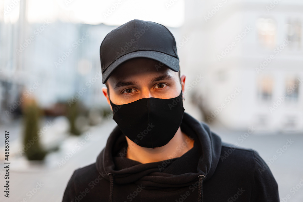 stylish modern portrait of handsome young guy with protective medical mask and black cap in fashion hoodie in the city. Urban male style and pandemic stylish modern portrait of handsome young guy with protective medical mask and black cap in fashion hoodie in the city. Urban male style and pandemic