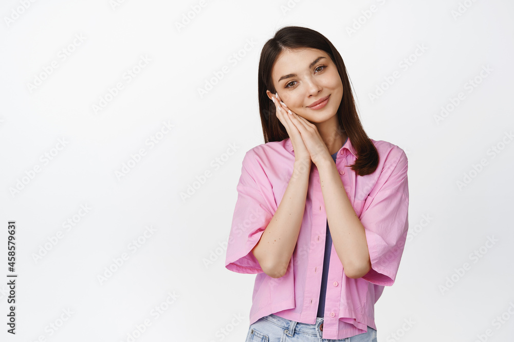 Cute young brunette woman with natural makeup, leaning face on hands as if sleeping, smiling tenderly, standing over white background Cute young brunette woman with natural makeup, leaning face on hands as if sleeping, smiling tenderly, standing over white background
