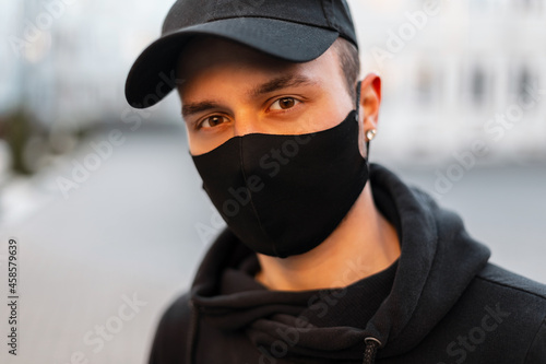 Urban fashion portrait of handsome young guy with protective medical mask and black cap in stylish casual hoodie outdoors. COVID-19