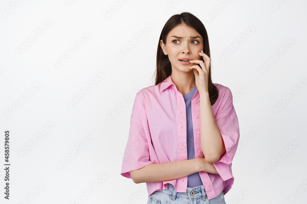 Anxious girl bites finger, looking scared and worried aside, overthinking, standing over white background Anxious girl bites finger, looking scared and worried aside, overthinking, standing over white background