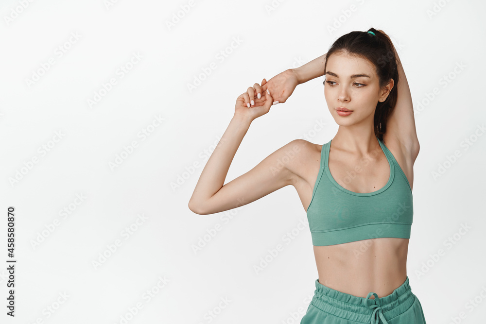 Young confident fitness girl stretching hands, workout warm-up in gym. Female athlete in sportsbra holding arm behind head, standing against white background Young confident fitness girl stretching hands, workout warm-up in gym. Female athlete in sportsbra holding arm behind head, standing against white background