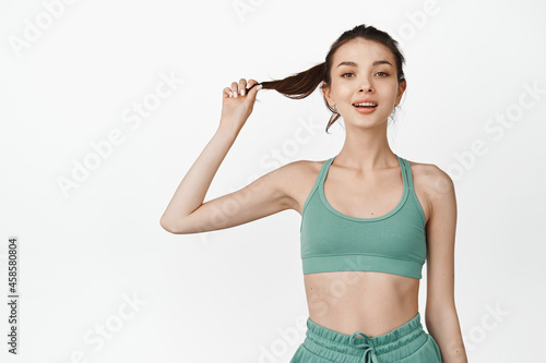 Slim and fit female athlete in sportsbra, looking confident and smiling at camera, standing over white background © Cookie Studio