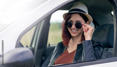 Young beautiful redhair woman in sunglasses smiling and looking at camera standing in car.