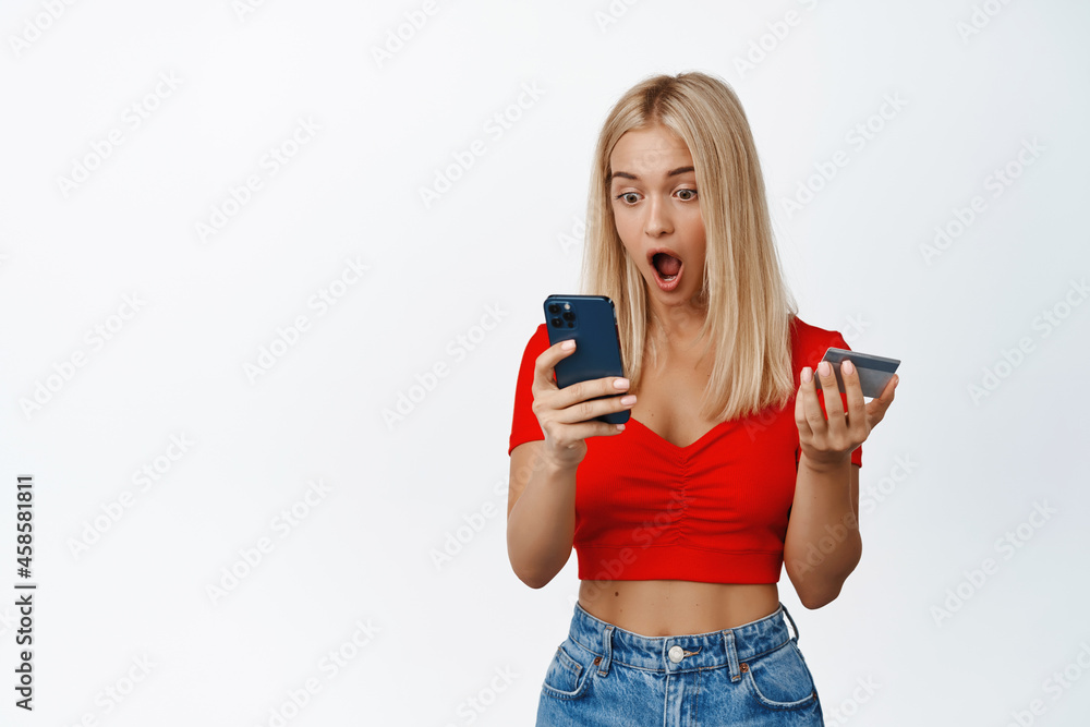 Surprised blond girl looking at smartphone screen while making order, browsing shopping site on mobile phone, holding credit card, white background Surprised blond girl looking at smartphone screen while making order, browsing shopping site on mobile phone, holding credit card, white background
