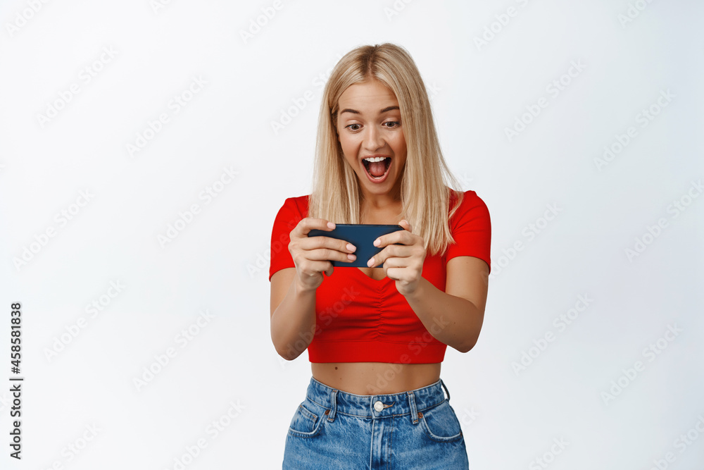 Happy blond woman playing video game on phone, watching on smartphone with amazed smiling face expression, white background Happy blond woman playing video game on phone, watching on smartphone with amazed smiling face expression, white background