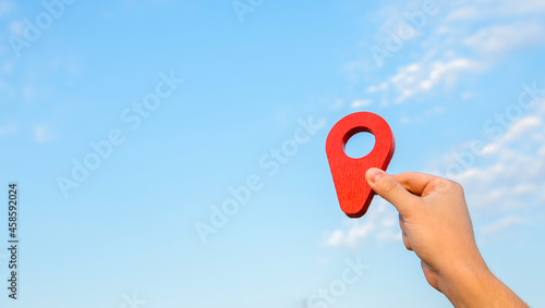 Red location pin in hand on sky background. The concept of location and orientation in space. Cartography. Tourism and travel. Land and real estate market. Air transportation and house moving.