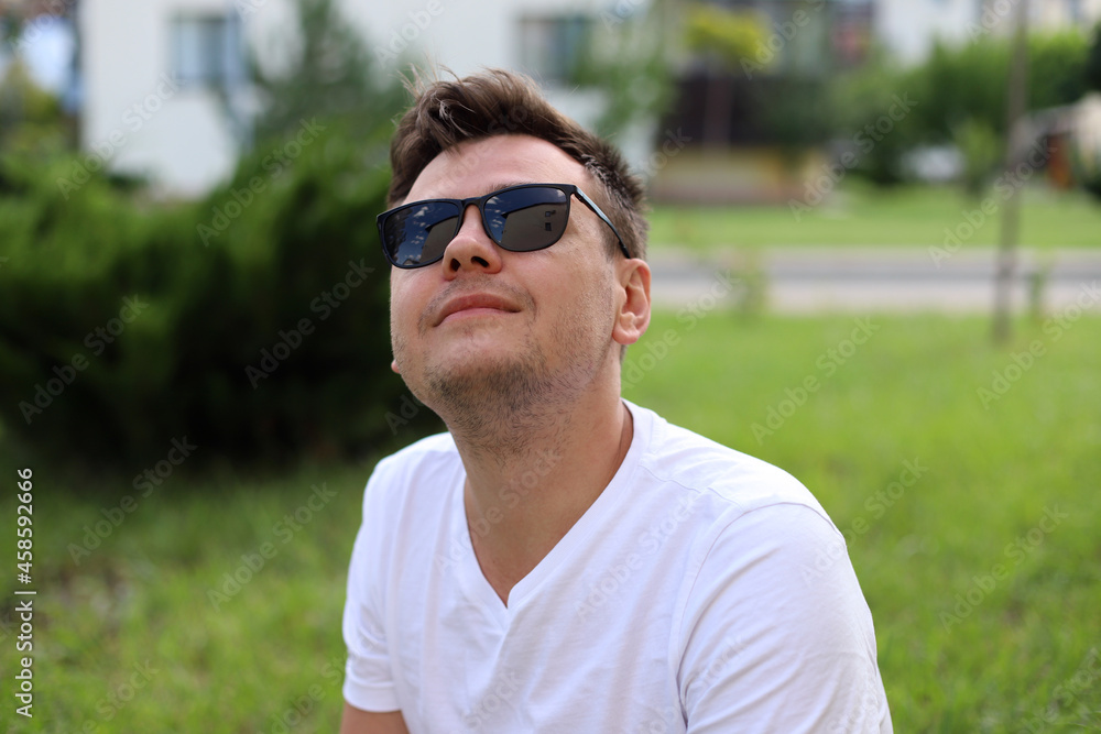 Smiling looking up young caucasian man with sunglasses in a white t-shirt on park with blurred sunny background. Selective focus. Portrait of a 32 year old young man. Smiling looking up young caucasian man with sunglasses in a white t-shirt on park with blurred sunny background. Selective focus. Portrait of a 32 year old young man.