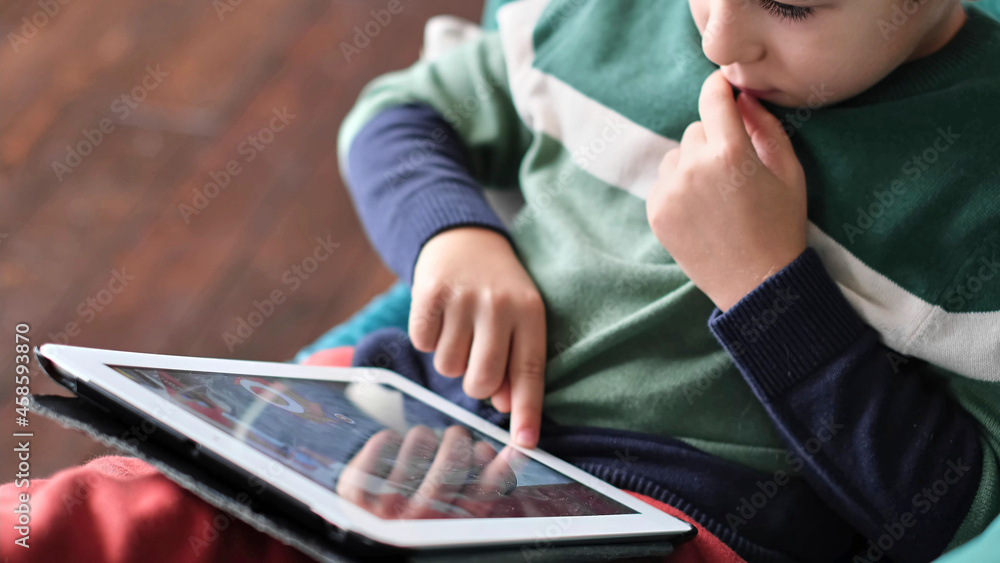 Cheerful little boy smiling while sitting on couch and using tablet at home. Modern kid and education technology. little boy looking at touch pad at home. Cheerful little boy smiling while sitting on couch and using tablet at home. Modern kid and education technology. little boy looking at touch pad at home.
