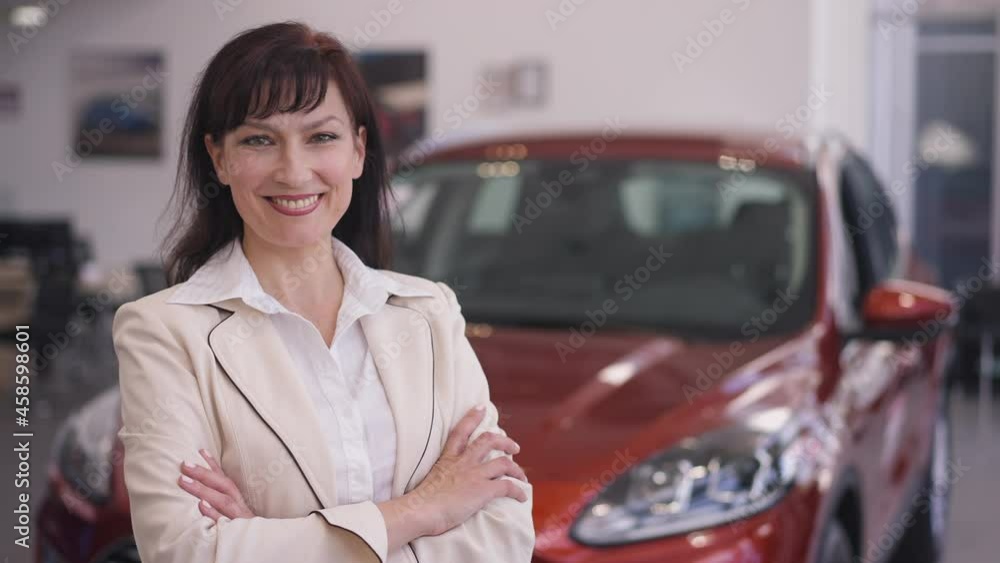Elegant confident beautiful woman crossing hands smiling looking at camera standing in car dealership indoors. Portrait of satisfied female buyer or professional dealer posing indoors in showroom Elegant confident beautiful woman crossing hands smiling looking at camera standing in car dealership indoors. Portrait of satisfied female buyer or professional dealer posing indoors in showroom