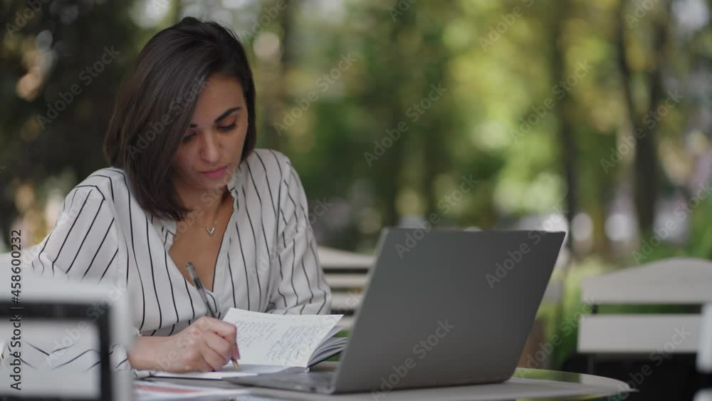 Business Woman Brunette Arabic Hispanic Ethnicity Works remotely while sitting in a summer cafe on a sunny day with a laptop and writes down with a pen and notebook Business Woman Brunette Arabic Hispanic Ethnicity Works remotely while sitting in a summer cafe on a sunny day with a laptop and writes down with a pen and notebook