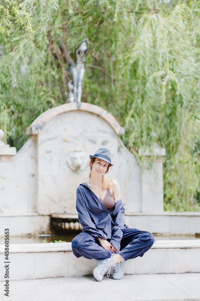 A casually dressed woman in a hat sits on the curb of a classic-style decorative park fountain A casually dressed woman in a hat sits on the curb of a classic-style decorative park fountain