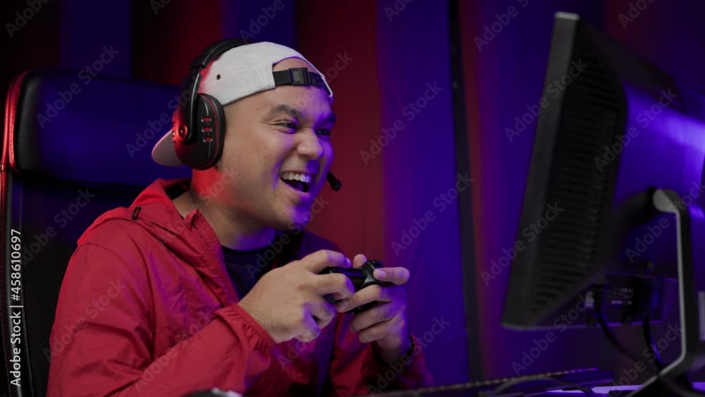 Excited and shocked face of Asian gamer with headphone holding joystick playing video game online sitting on chair at living room. Indian professional gamer streaming on social playing game very fun Excited and shocked face of Asian gamer with headphone holding joystick playing video game online sitting on chair at living room. Indian professional gamer streaming on social playing game very fun