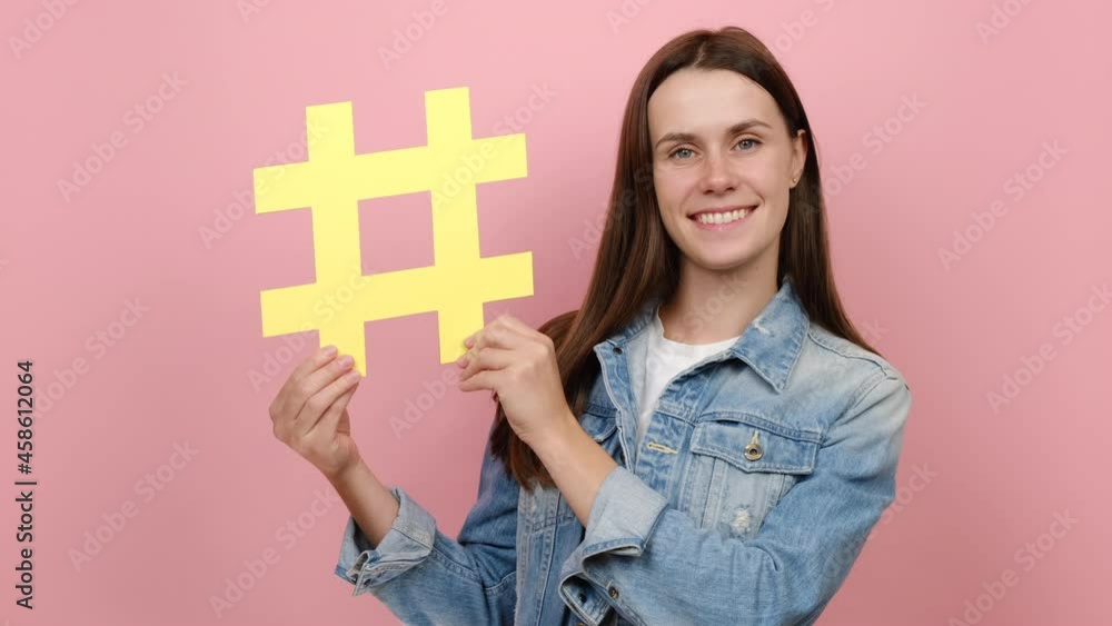 Happiness sweet young female 20s with toothy smile demonstrate hashtag symbol, positive looking at camera, tagging famous blog, viral content, wears denim jacket, isolated over pink studio background Happiness sweet young female 20s with toothy smile demonstrate hashtag symbol, positive looking at camera, tagging famous blog, viral content, wears denim jacket, isolated over pink studio background