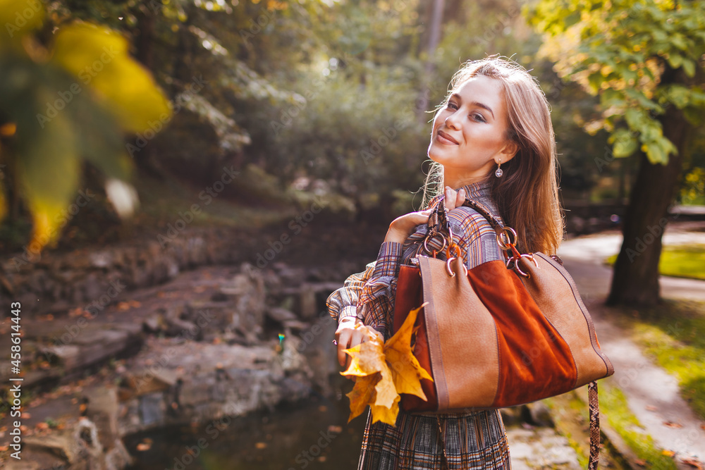 Portrait of woman holding brown handbag in fall park. Stylish girl wears trendy accessories and clothes walking outdoors Portrait of woman holding brown handbag in fall park. Stylish girl wears trendy accessories and clothes walking outdoors