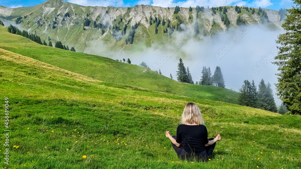 A blond woman meditating on green grass, looking over a beautiful scenery on the valley, mountains, clouds on a bright sunny day. A blond woman meditating on green grass, looking over a beautiful scenery on the valley, mountains, clouds on a bright sunny day.