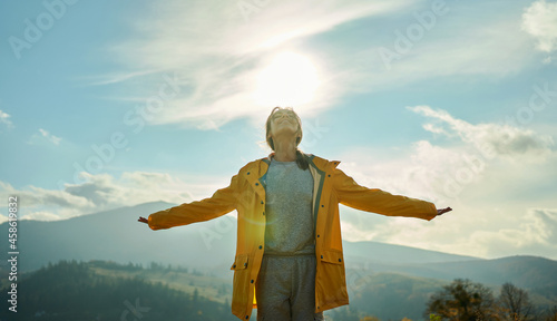 Joyful freedom girl in yellow jacket standing with open arms around mountains and hills, sun lights over hand, feeling happiness