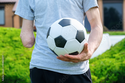 Full length young caucasian man with amputated arm, holds of soccer ball while standing on the lawn in the backyard of his home, disability rehabilitation and active lifestyle