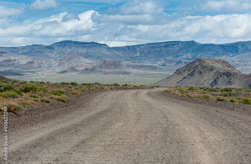 High Desert, Nevada, USA - May 17, 2011: Gray durt abd pebbles road meanders though wide landscape with mountain range in back under blue cloudscape. Green shrub around.