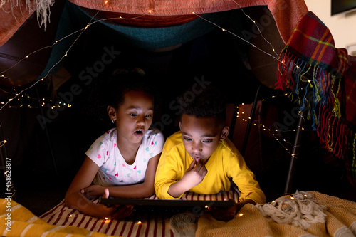 Surprised african american siblings lying in makeshift tent, using tablet