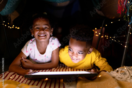 Happy african american siblings lying in makeshift tent, using tablet