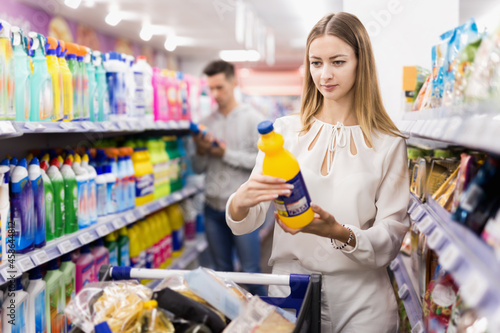 Portrait of young glad positive woman with shopping cart choosing household chemicals in supermarket