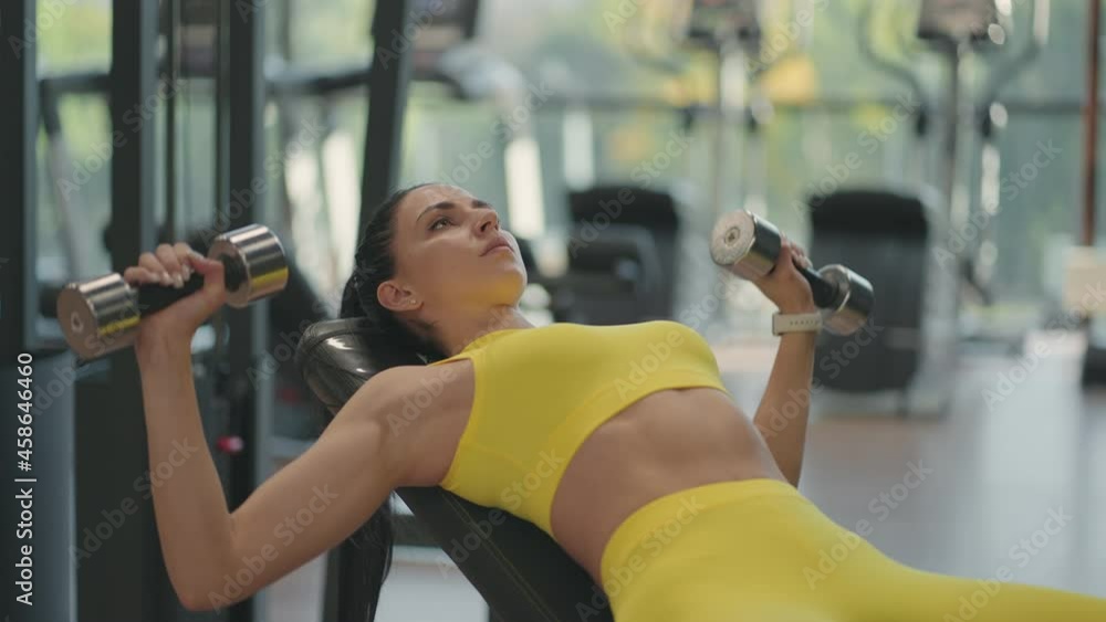A young brunette woman in a yellow suit is pushing a dumbbell while lying on the bench. Dumbbell bench press. Exercise for chest muscles A young brunette woman in a yellow suit is pushing a dumbbell while lying on the bench. Dumbbell bench press. Exercise for chest muscles