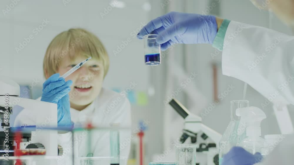 Two boys preparing scientific experiment with teacher. Laboratory interior. Passing multi colored liquids with pipettes, using glass tubes Two boys preparing scientific experiment with teacher. Laboratory interior. Passing multi colored liquids with pipettes, using glass tubes