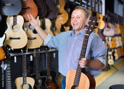 Adult glad smiling guitarist is standing with acoustic guitar in music store.