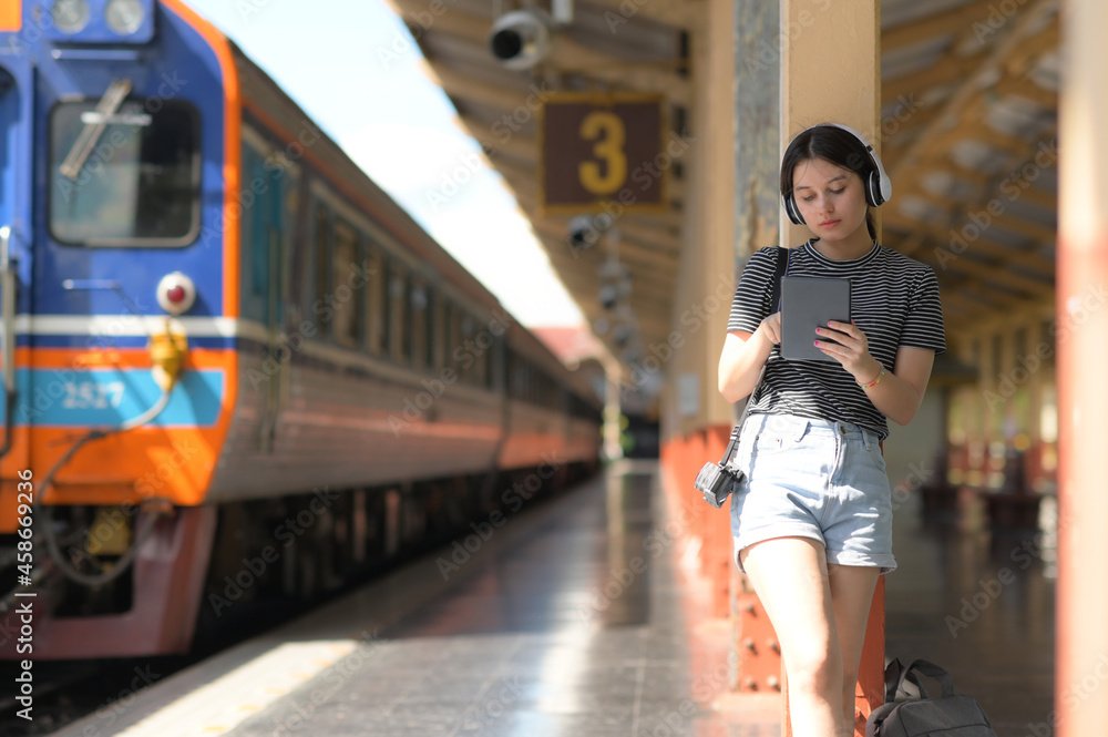 A teenage woman wearing headphones listening to music from an app on her tablet while waiting for a train..