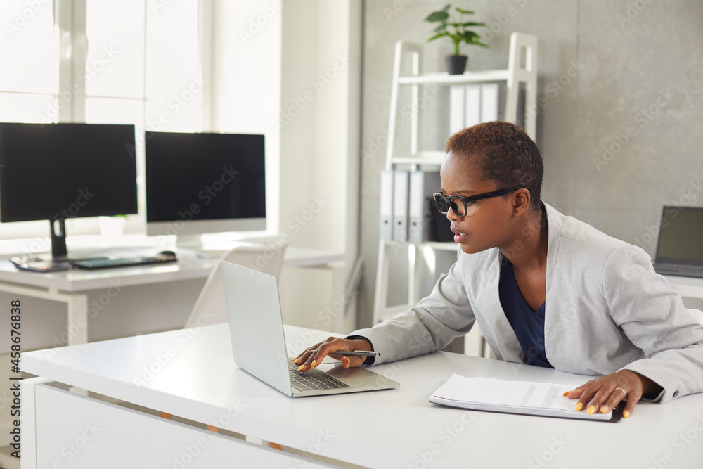 Millennial African American woman sit at desk in office work online on laptop consider financial business paperwork. Young biracial businesswoman use computer at workplace consult client customer.