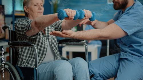 Close up of disabled woman using dumbbells for recovery therapy in nursing home. Medical assistant helping retired woman in wheelchair use objects for physical exercise and activity