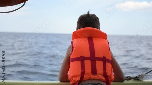 little child in orange safe lifejacket during summer leisure activity in sea on boat board with other vessel sailing near photo