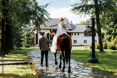 Wedding ceremony in villa outdoors, newlywed couple look at each other in front of country house, bride in white dress sitting on brown horse, groom in suit holds reins, back view. Just married