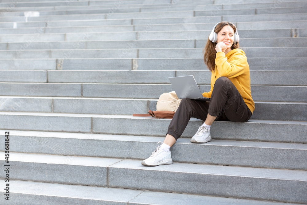 Student life, additional education. Adult caucasian woman in casual wear and headset sits on the steps of the stairs with a laptop outdoor and ponder something, selected focus.