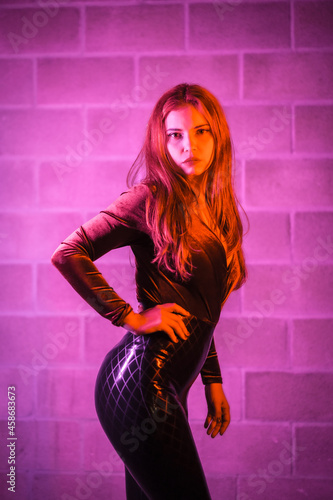 Red-haired girl in latex, brown and black tight suit on a brick wall, posed at night illuminated with pink light photo