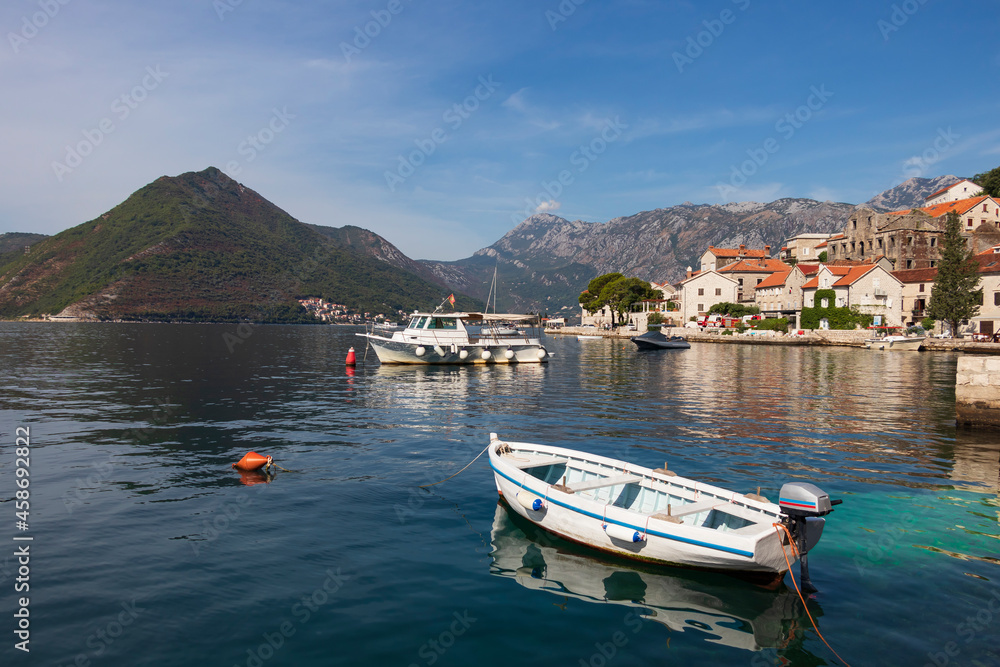 A picturesque seascape on the Bay of Kotor, mountains and the Perast embankment, in the foreground a fishing motor boat is moored, the silhouette of which is reflected on the calm surface of the water
