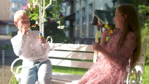 Caucasian girl and boy brother and sister blowing bubbles in park with parents watching from behind. Family lifestyle, outdoors activities, summer holidays with kids. 